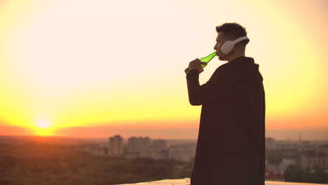 A-man-drinks-beer-standing-on-the-roof-and-contemplates-the-world.-Look-at-the-view-of-the-city-at-sunset-from-a-height.-The-roof-of-a-skyscraper.